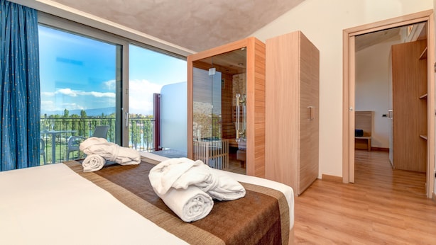 A modern hotel room with a large bed, a glass shower enclosure, and a balcony with a view of the city. - Hotel Rivus (Peschiera del Garda)