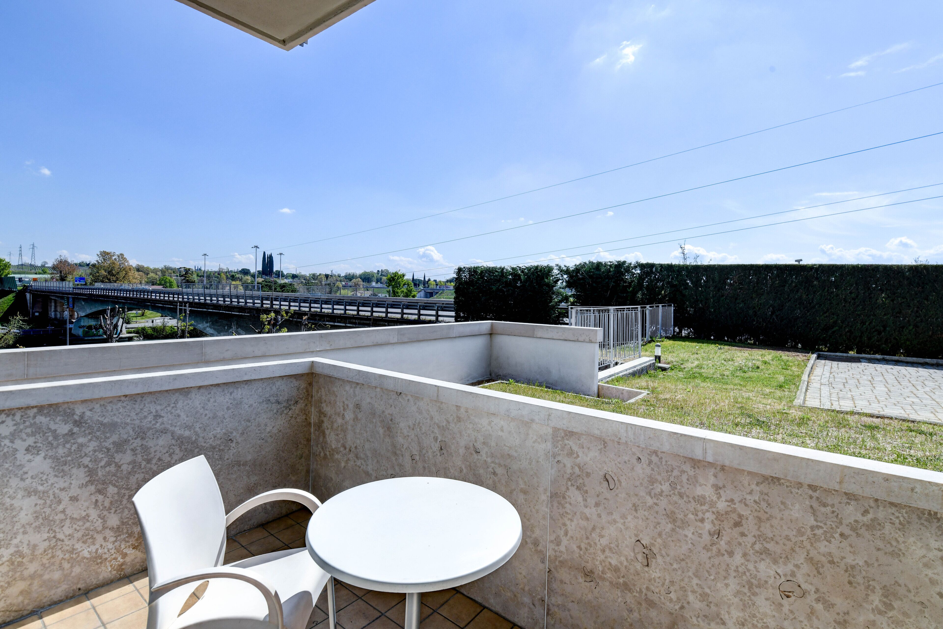 A balcony with a white table and chairs, a view of a bridge, and a hedge.