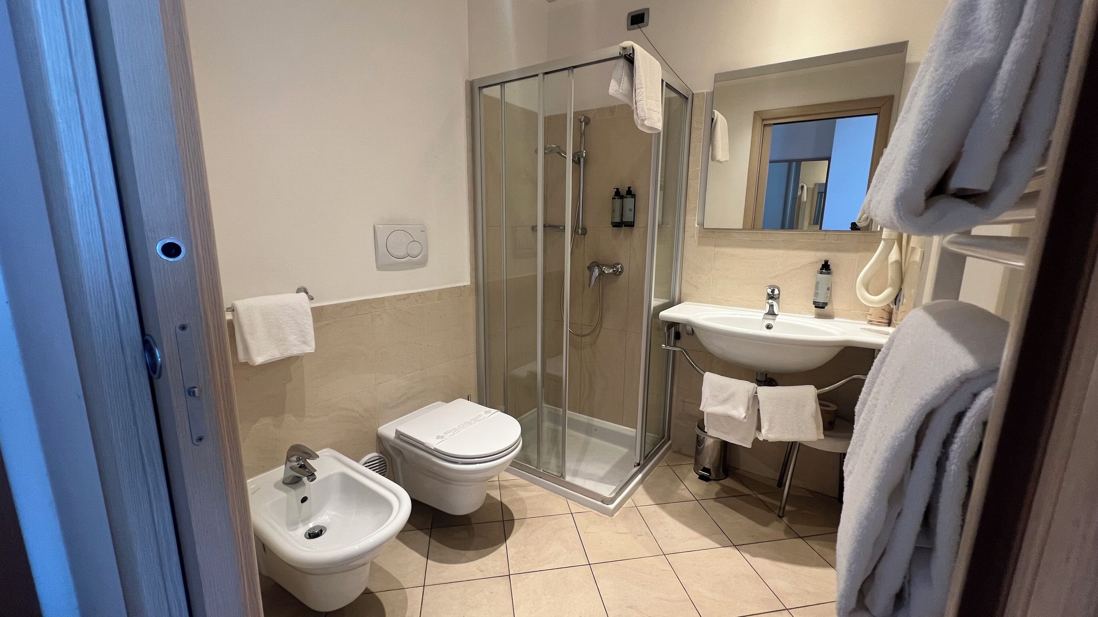 A bathroom with a glass shower enclosure, a bidet, a toilet, and a sink with a mirror above.