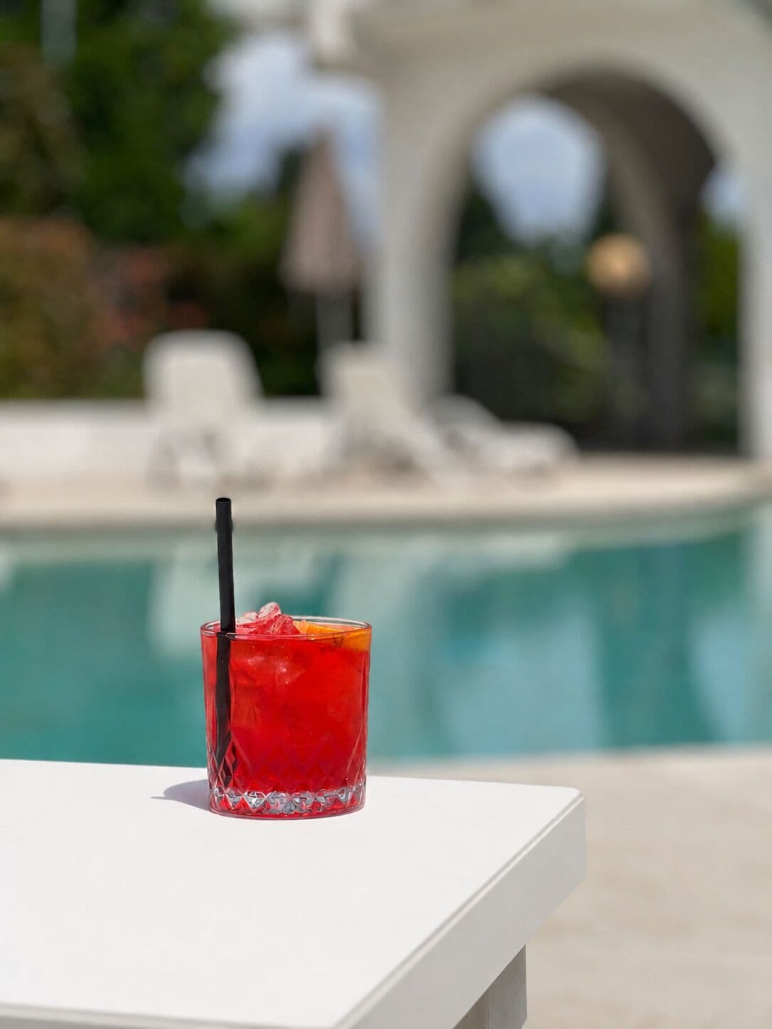 A glass of red drink with a straw on a white table by a pool.