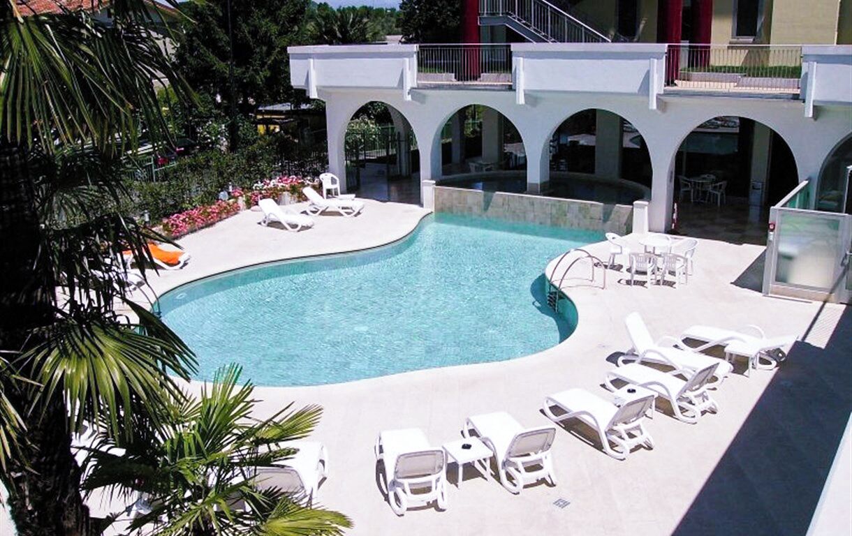 A swimming pool with lounge chairs, a covered area with arches, and a building with balconies.