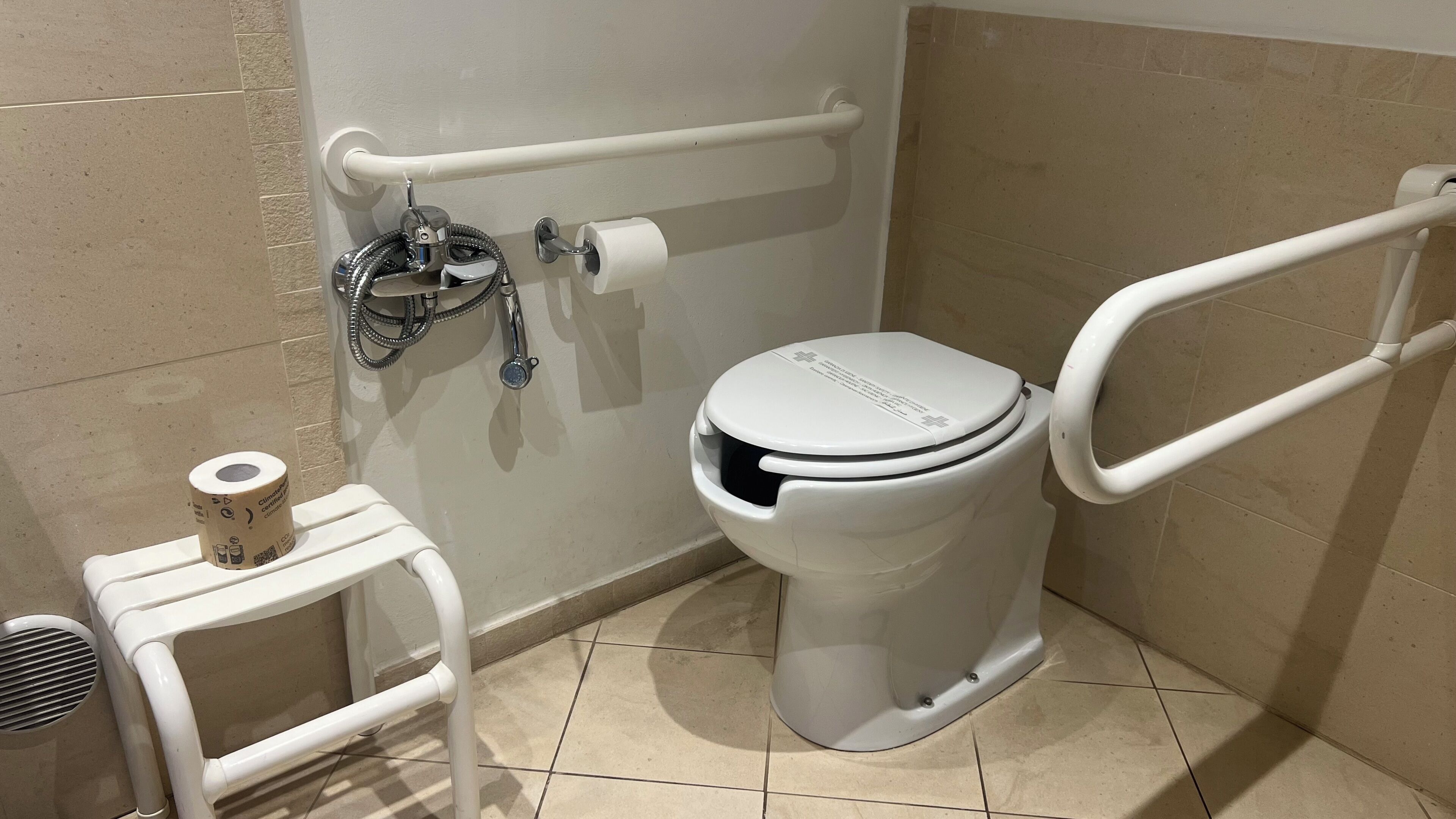 A bathroom with a white toilet, a handrail, a grab bar, a toilet paper holder, and a vent.