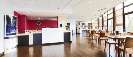 A spacious hotel lobby with a reception desk, wooden flooring, and large windows offering natural light.