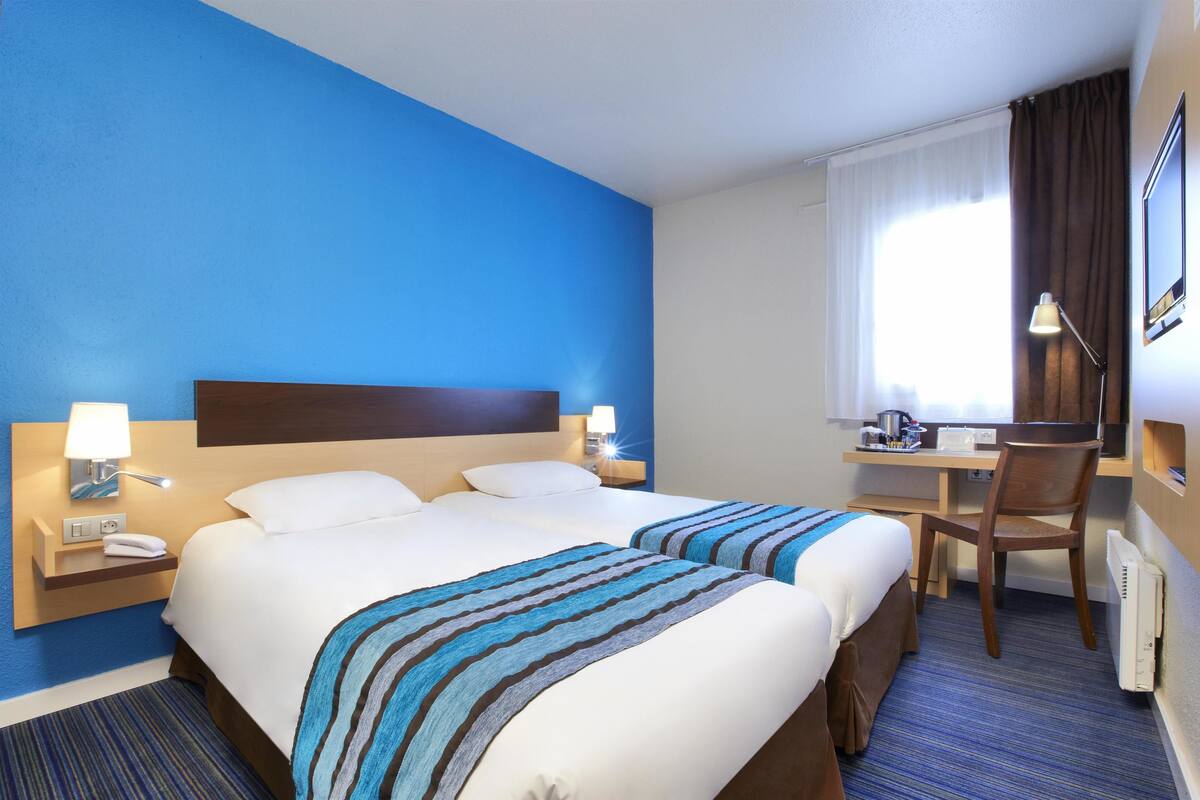 A hotel room with two beds, a desk, a chair, and a window with curtains.