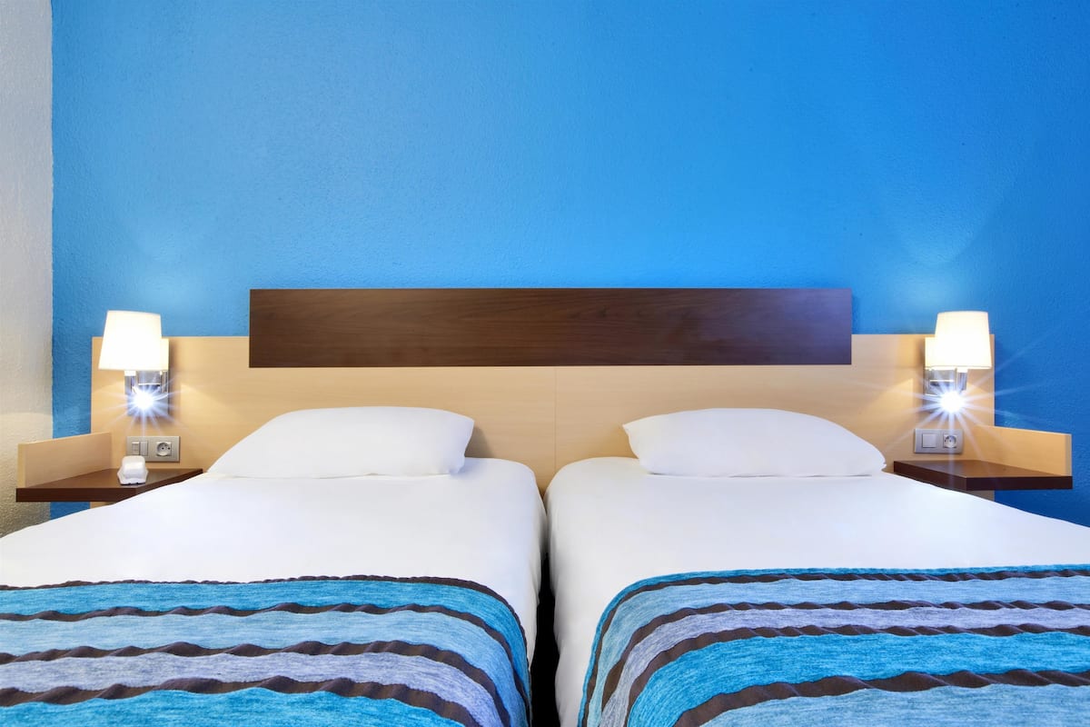 A hotel room with two beds, each with a wooden headboard, white bedding, and a blue-striped blanket. There are bedside lamps on each nightstand.