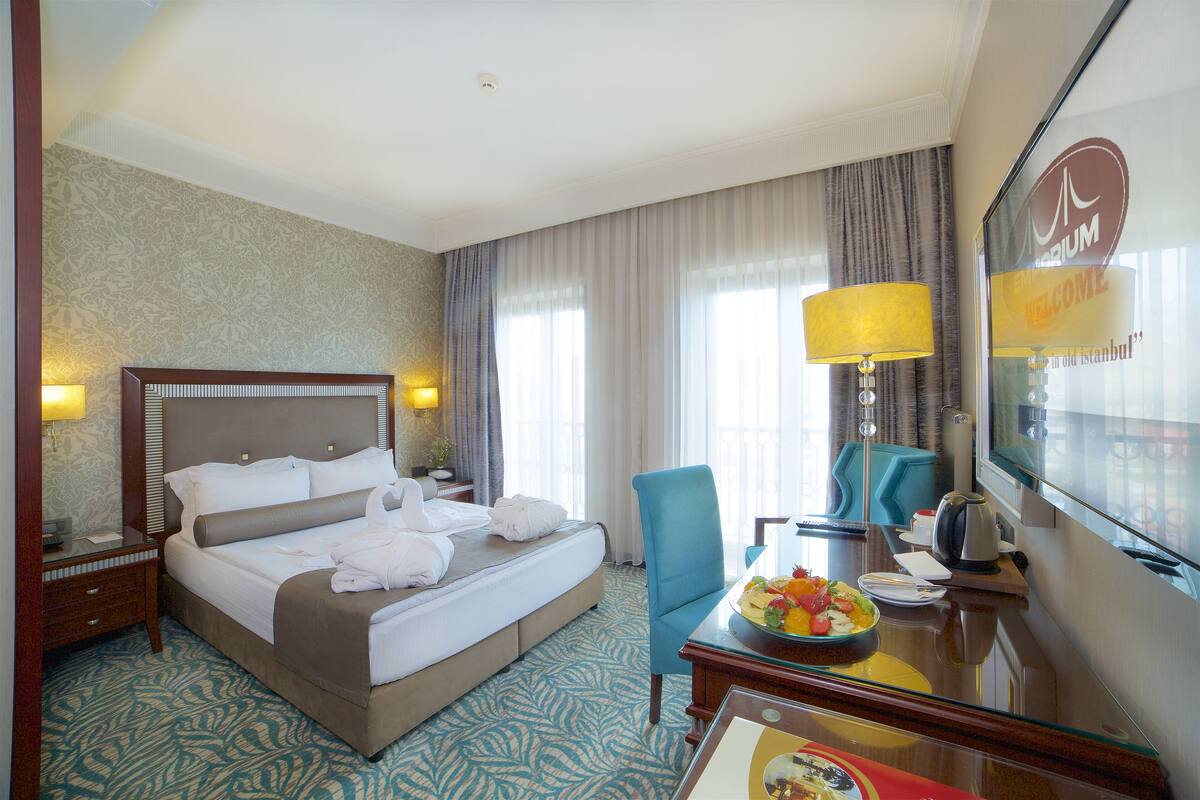 A hotel room with a large bed, a desk with a lamp, a chair, and a TV.