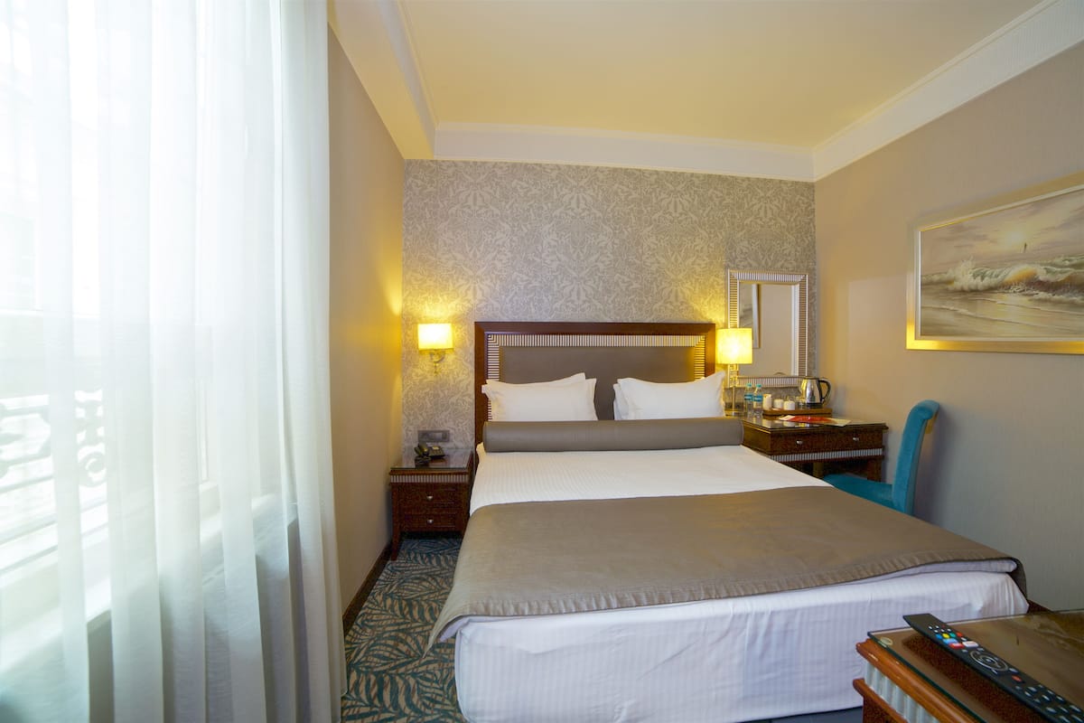 A hotel room with a large bed, a nightstand with a lamp, a desk with a chair, and a painting on the wall.
