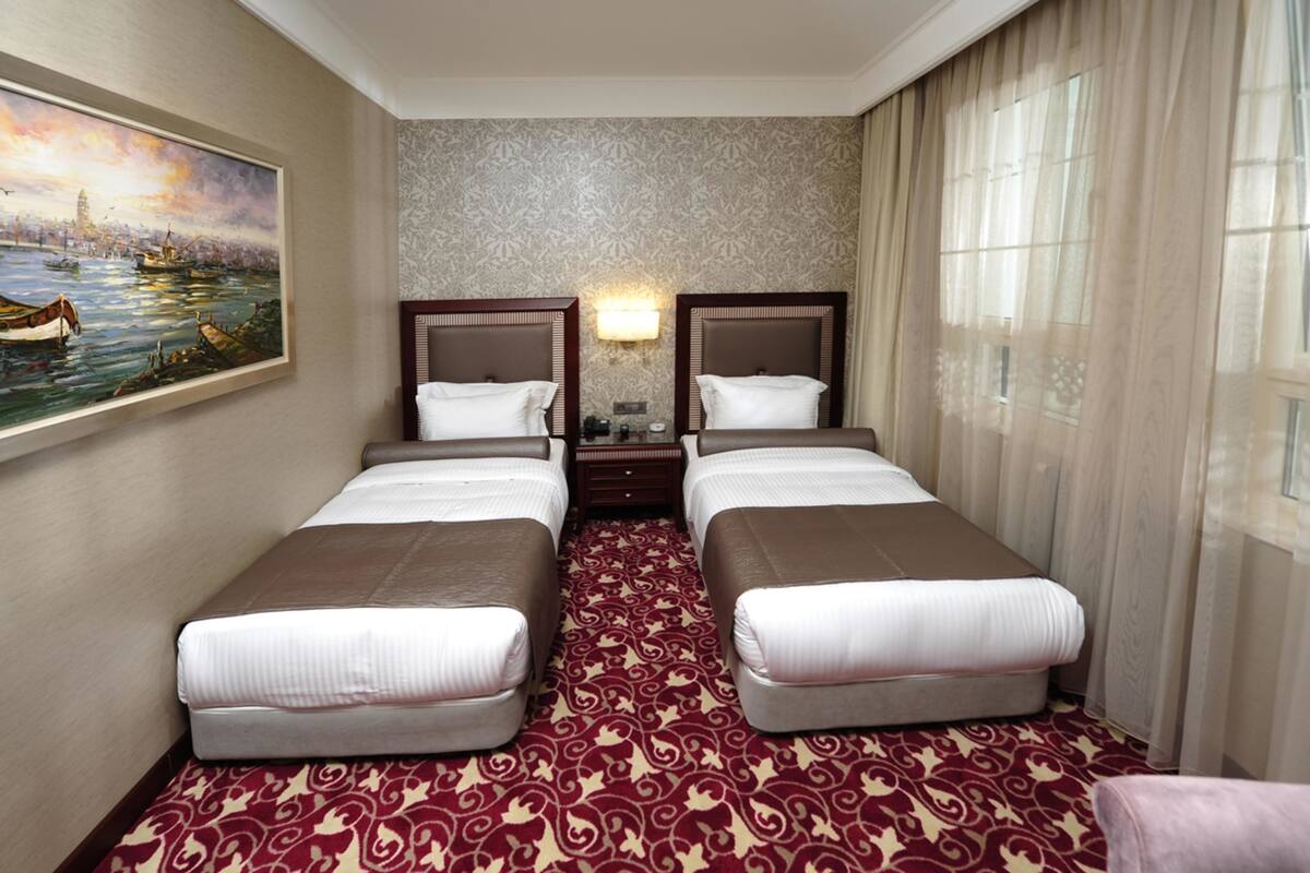 A hotel room with two beds, a patterned carpet, a painting on the wall, and a window with curtains.