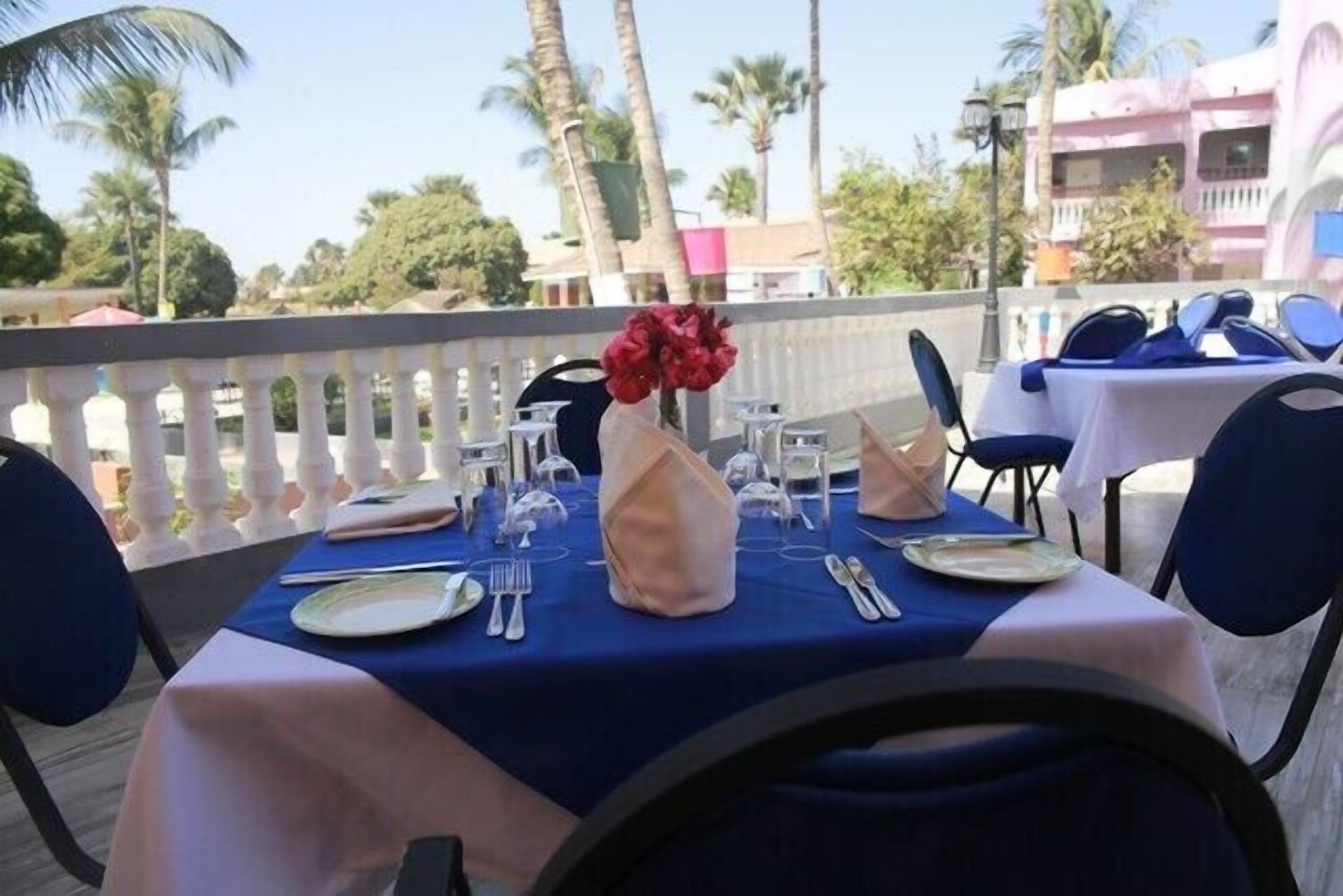 Un espacio de comedor al aire libre con mesas preparadas para dos personas, con platos blancos, cubiertos y un florero con flores rojas.