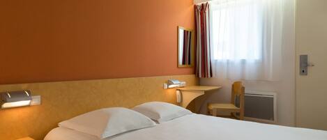 A hotel room with a bed, a bedside table with a lamp, a chair, a window with curtains, and a wall-mounted phone.