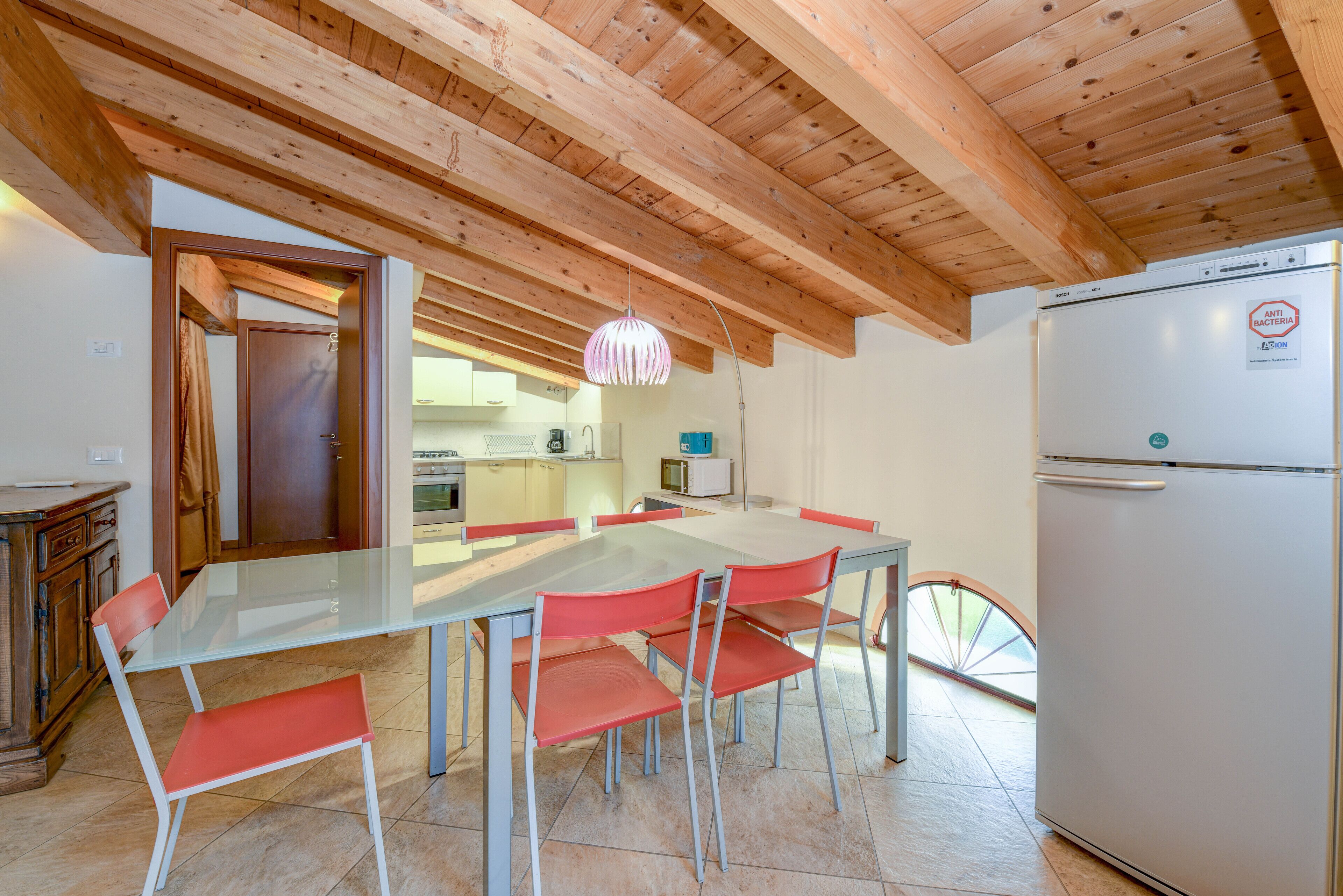 Una cocina moderna con techo de madera, una mesa de comedor de vidrio con sillas rojas, un refrigerador blanco y una ventana con vistas al exterior.