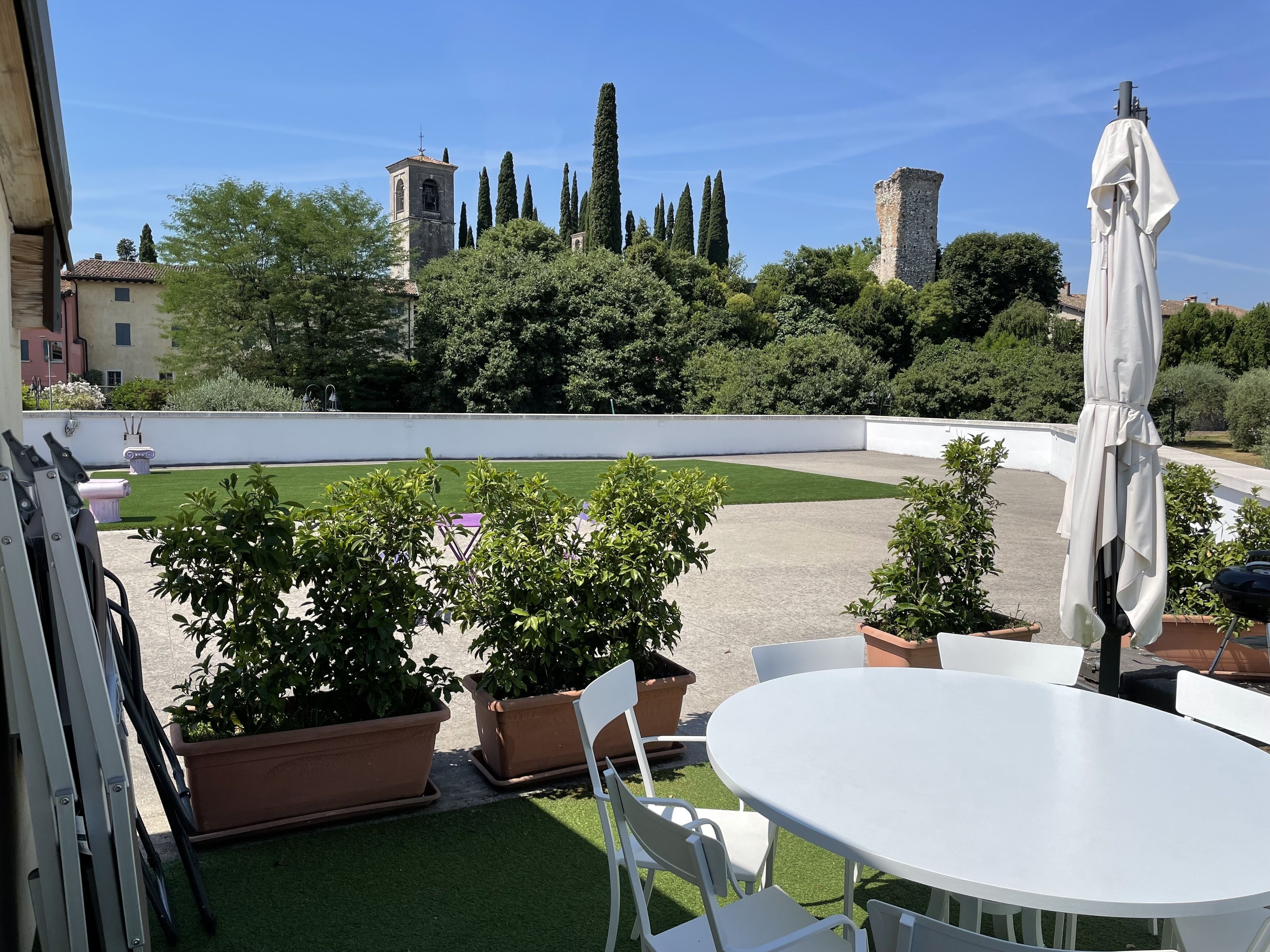 A rooftop terrace with a white table and chairs, surrounded by potted plants and a small lawn.
