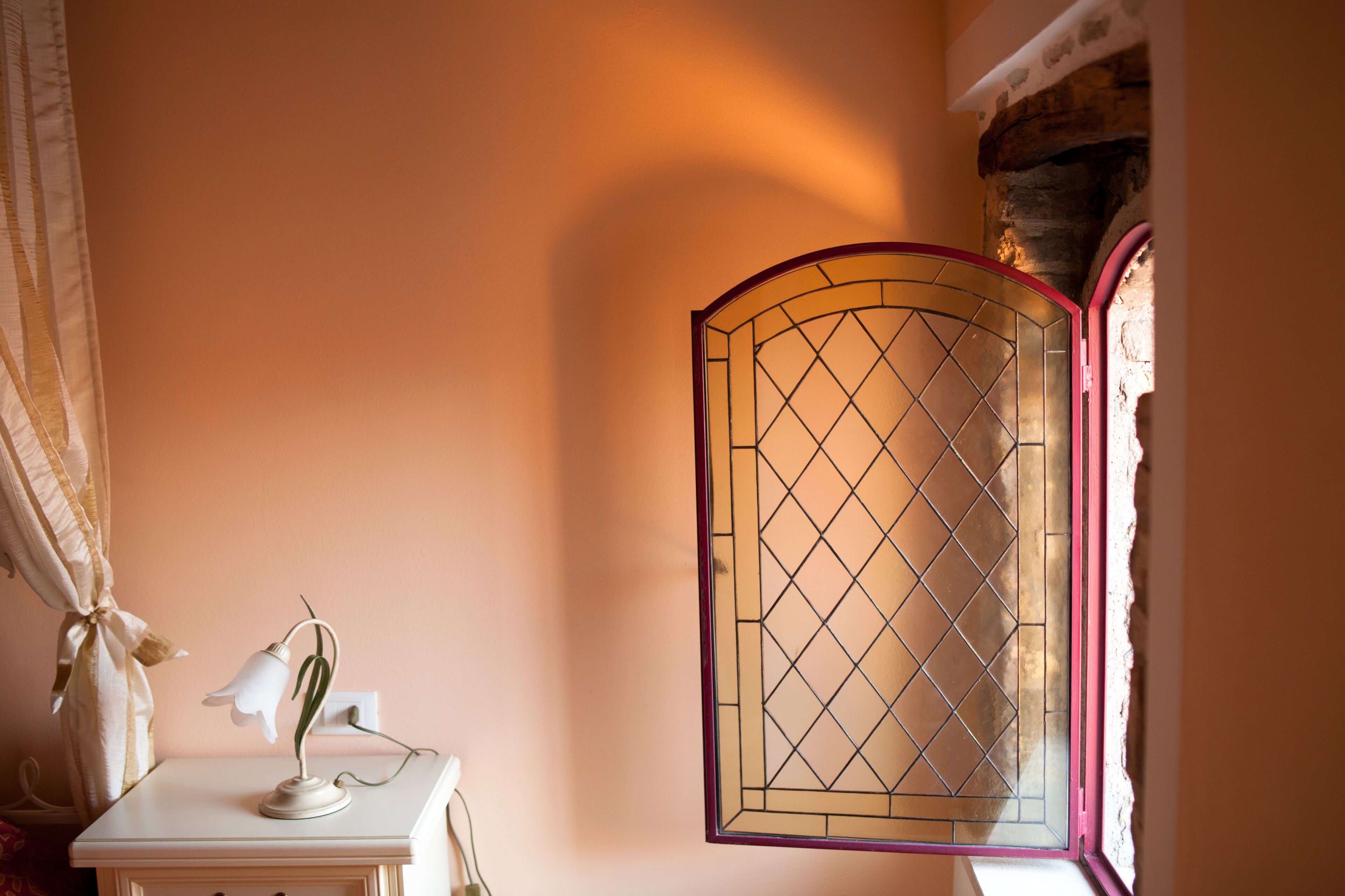 A room with a white bedside table, a lamp, and a window with a decorative glass panel.
