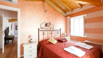 A bedroom with a wooden ceiling, a bed with a red bedspread, a bedside table with a lamp, and a window with blinds.