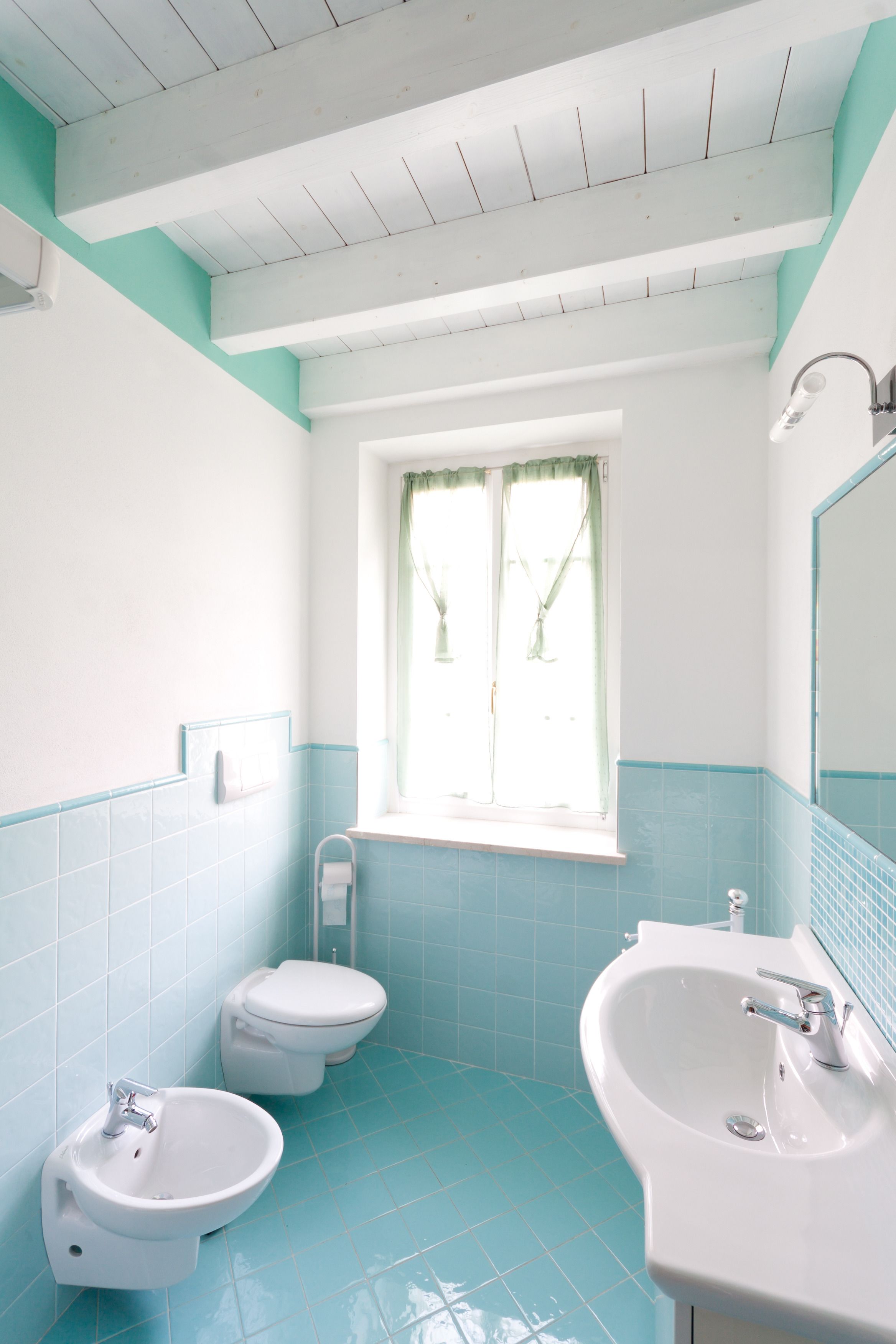 Egy fürdőszoba, amelyben WC, bidé és mosdókagyló található, türkiz és fehér csempékkel, valamint függönnyel ellátott ablakkal.