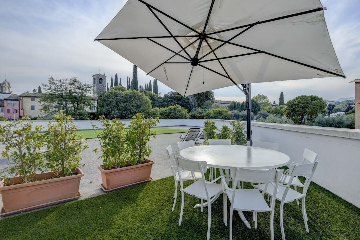 A rooftop terrace with a round table and chairs, surrounded by potted plants and an umbrella.