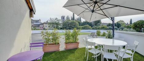 A rooftop terrace with a white table and chairs, a purple folding chair, and a large umbrella. There are potted plants and a view of buildings and trees in the background.