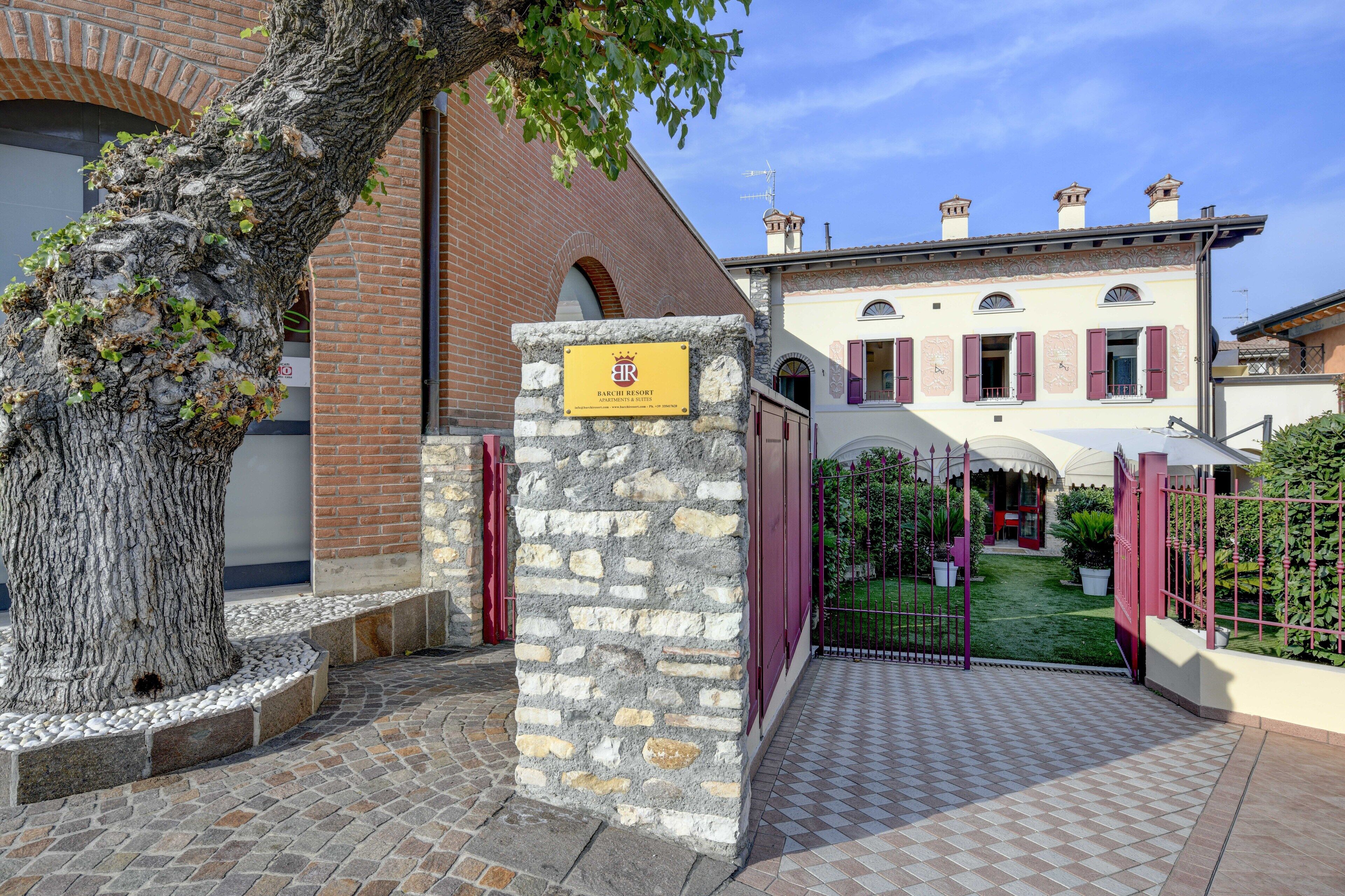 A stone pillar with a plaque, a brick building, and a gated entrance with a small garden.