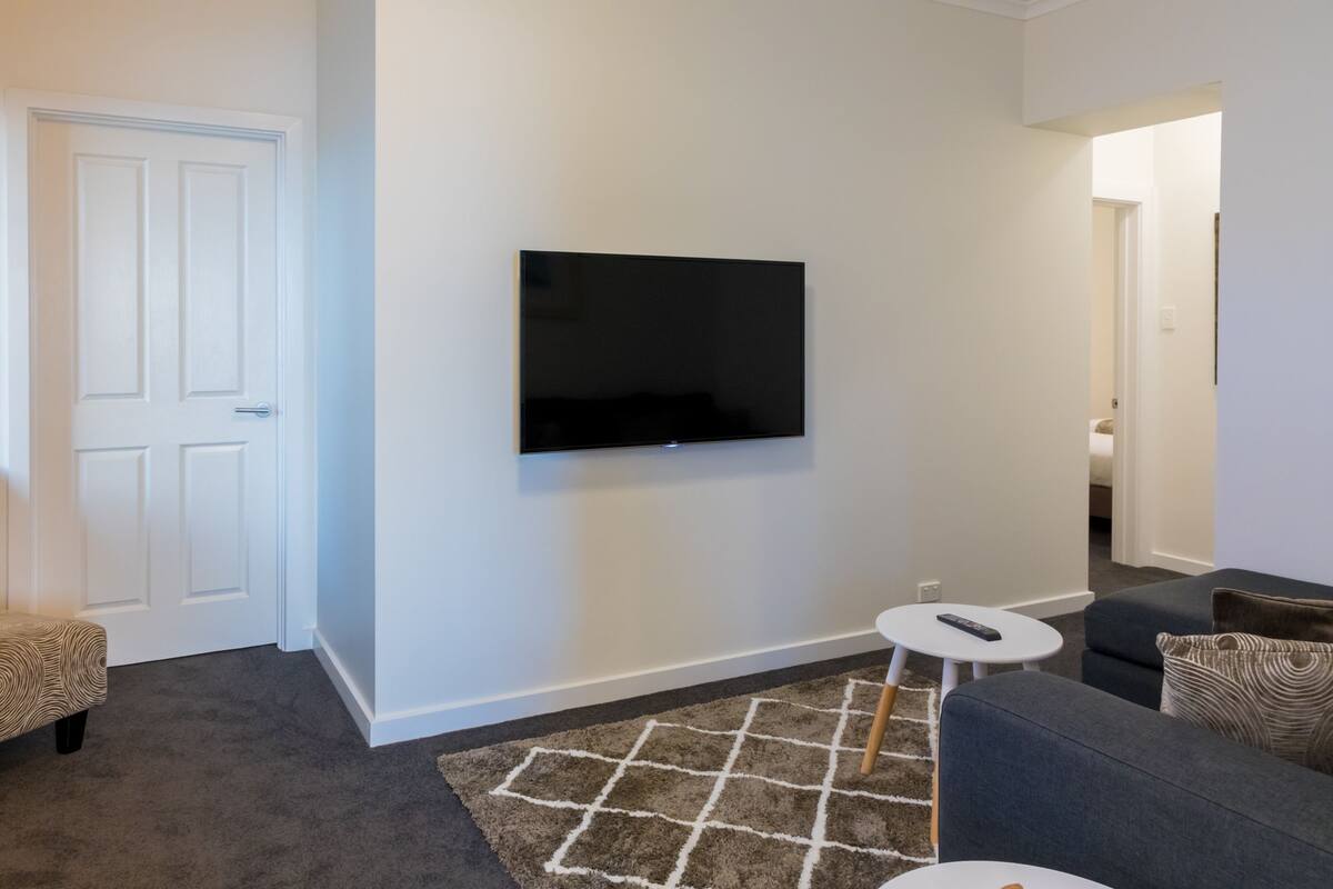 A living room with a flat-screen TV mounted on the wall, a grey sofa, a small white side table, and a patterned rug.