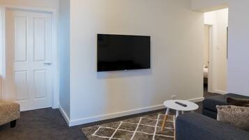 Uma sala de estar com televisão de tela plana na parede, um sofá cinza, uma mesinha lateral branca e um tapete estampado.