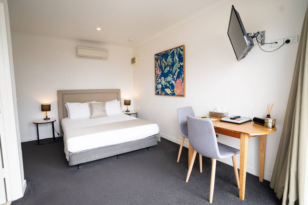 A hotel room with a bed, bedside tables, a desk, a chair, a flat-screen TV mounted on the wall, and a floral artwork.