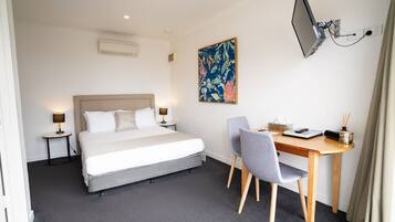 A hotel room with a bed, bedside tables, a desk, a chair, a flat-screen TV mounted on the wall, and a floral artwork.