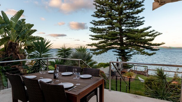 A wooden table set for dining with chairs, overlooking a body of water and a mountainous landscape. - Bay 10 Accommodation (Port Lincoln)