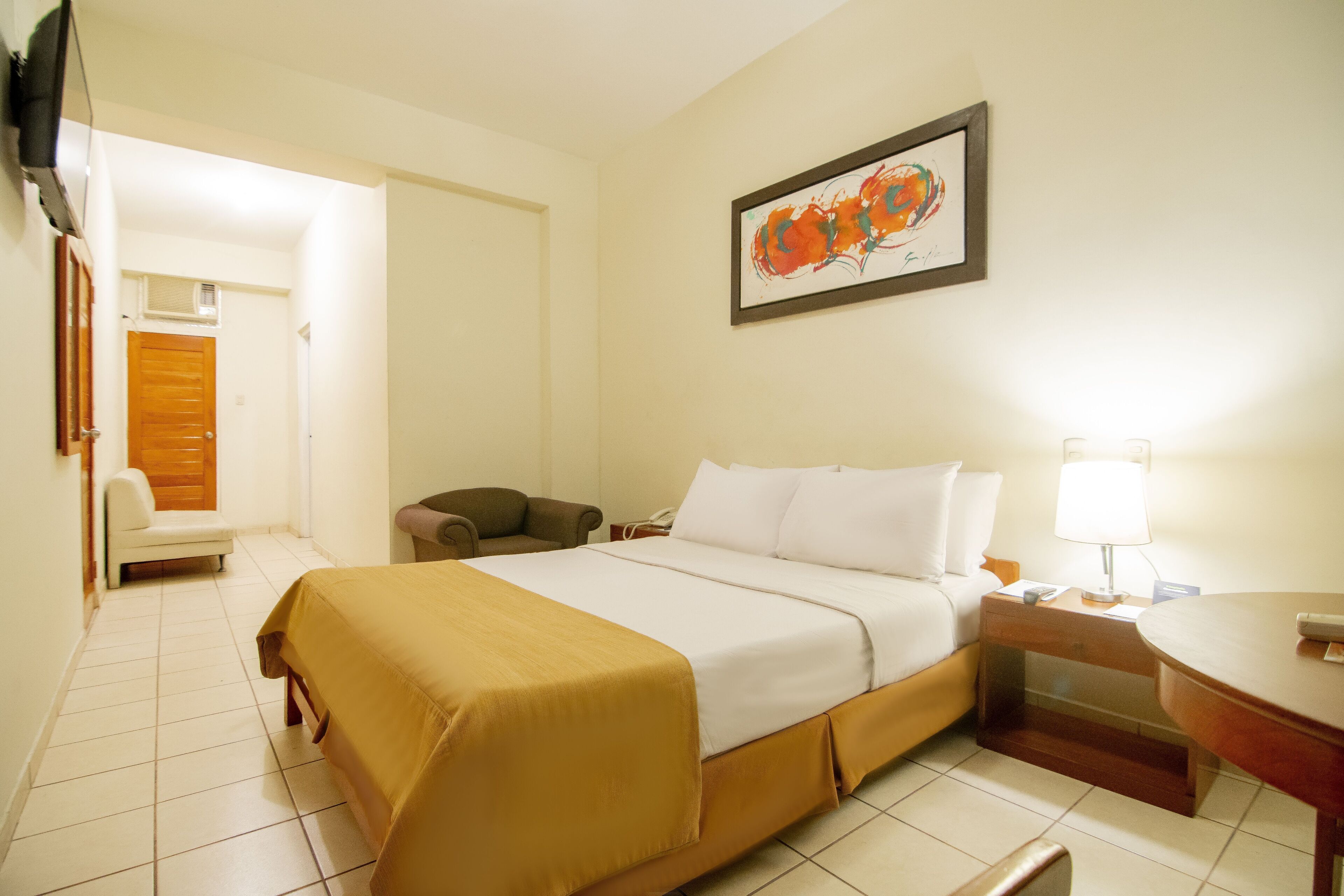 A hotel room with a bed, a chair, a desk with a lamp, and a painting on the wall.