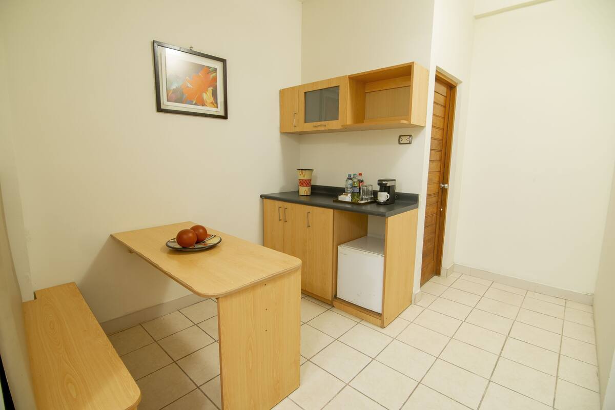 Una pequeña cocina americana con una mesa de madera, un refrigerador y una encimera con una cafetera y otros objetos.
