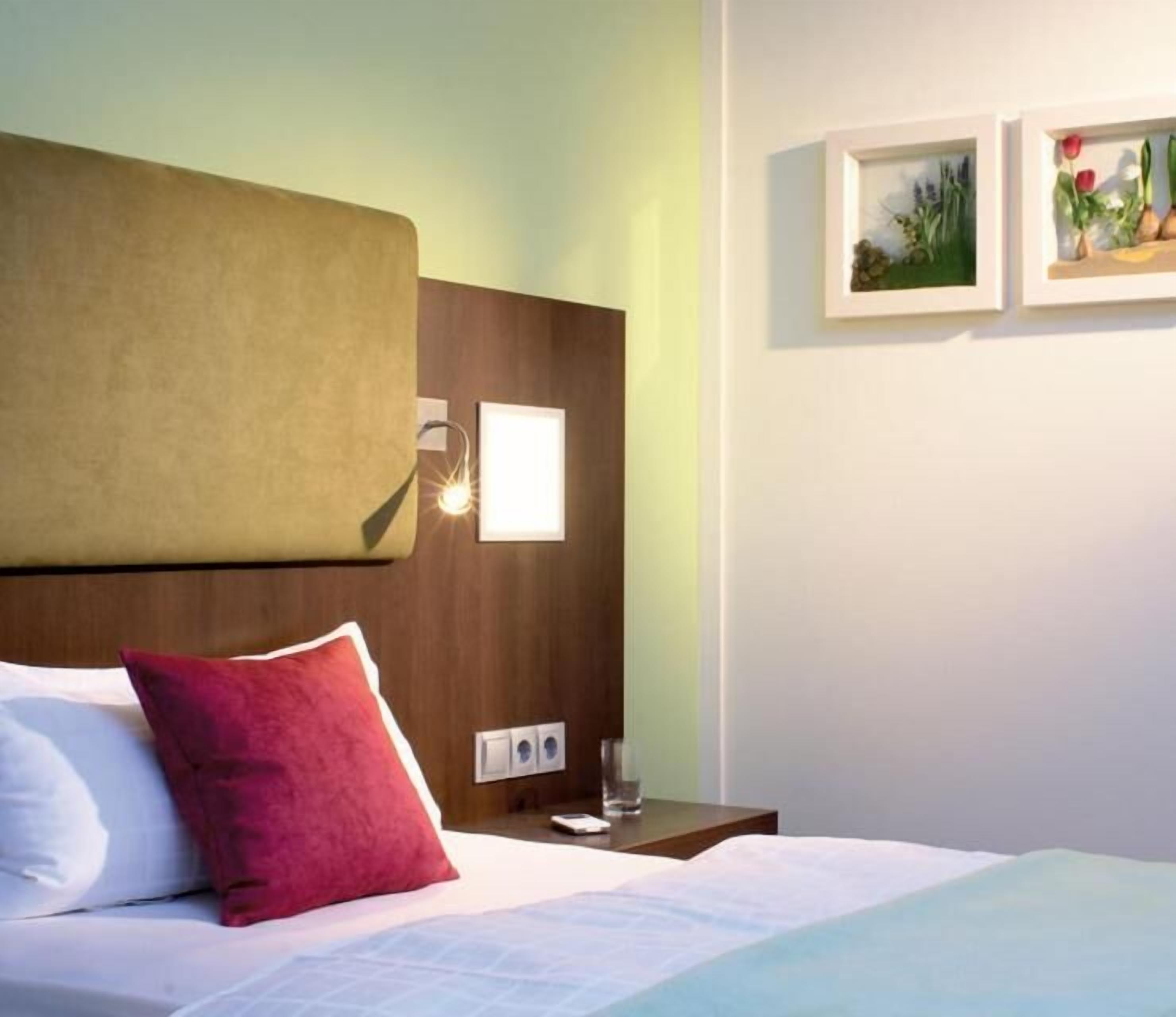 A hotel room with a bed, a nightstand, a wall-mounted lamp, and two framed pictures.