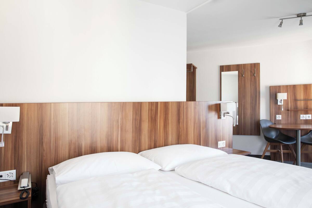 Habitación de hotel con cabecera de madera, cama con ropa blanca, escritorio con silla y espejo.