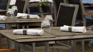 Esstische im Freien mit Stühlen, einige mit gefalteten Servietten und Gläsern, unter Sonnenschirmen.