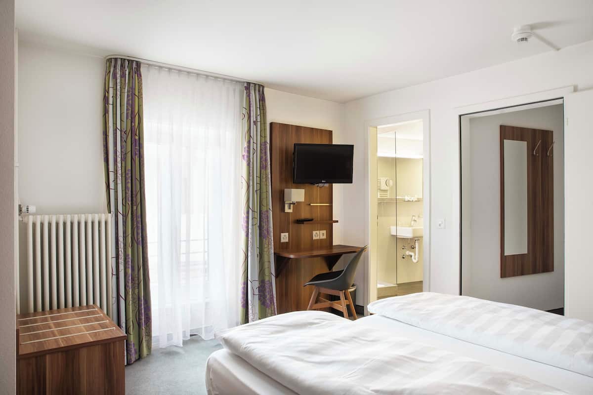 Habitación de hotel con cama, escritorio, silla, televisor, radiador y baño visible a través de una puerta abierta.