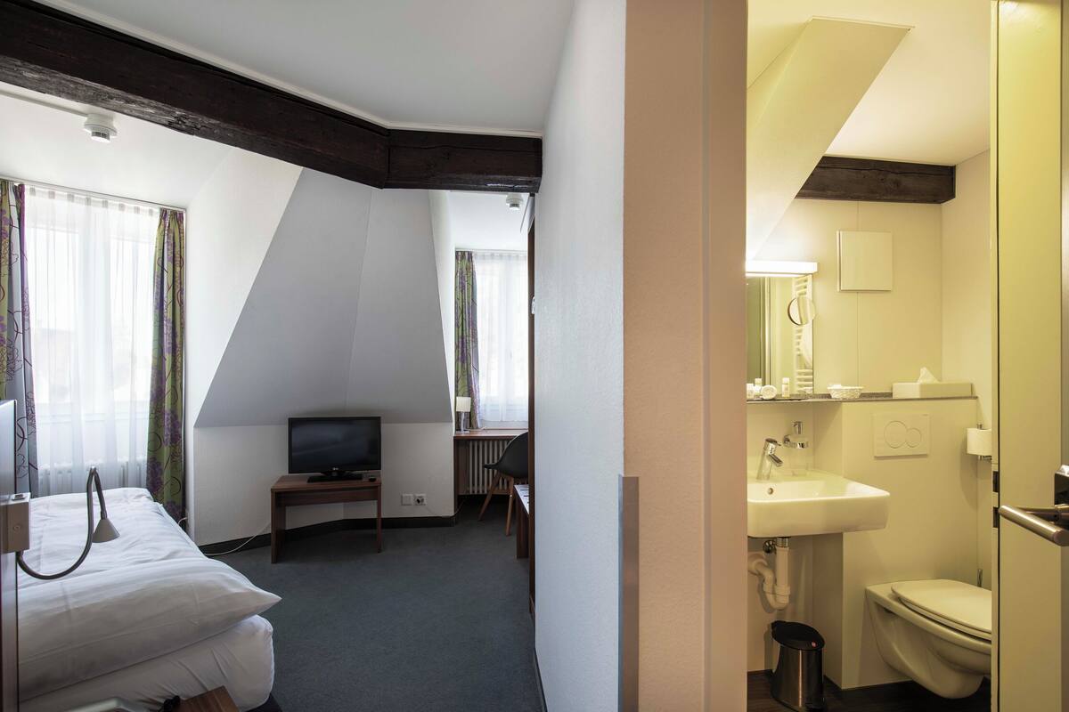 Habitación de hotel con cama, escritorio con televisor, ventana con cortinas y baño con lavamanos e inodoro.