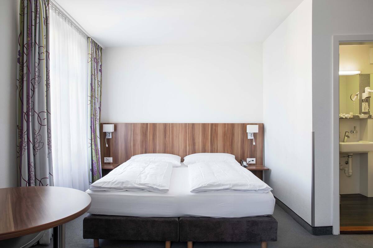 Habitación de hotel con cama, mesitas de noche, una pequeña mesa de madera y vistas al baño.