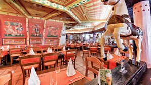 Ein Restaurant mit Holztischen und -stühlen, einer großen Pferdestatue und farbenfroher Deckenverzierung.