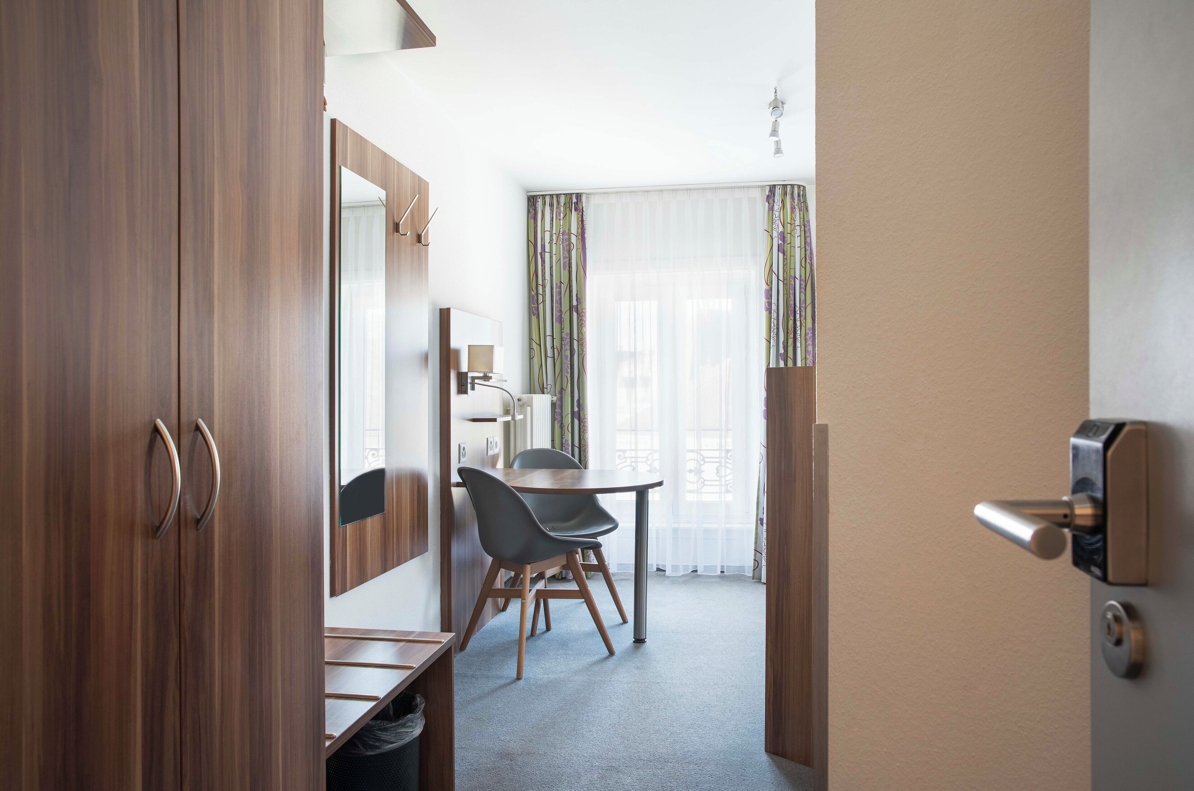 Una habitación de hotel moderna con un armario de madera, una pequeña mesa de comedor con sillas, un espejo grande y una ventana con cortinas translúcidas.