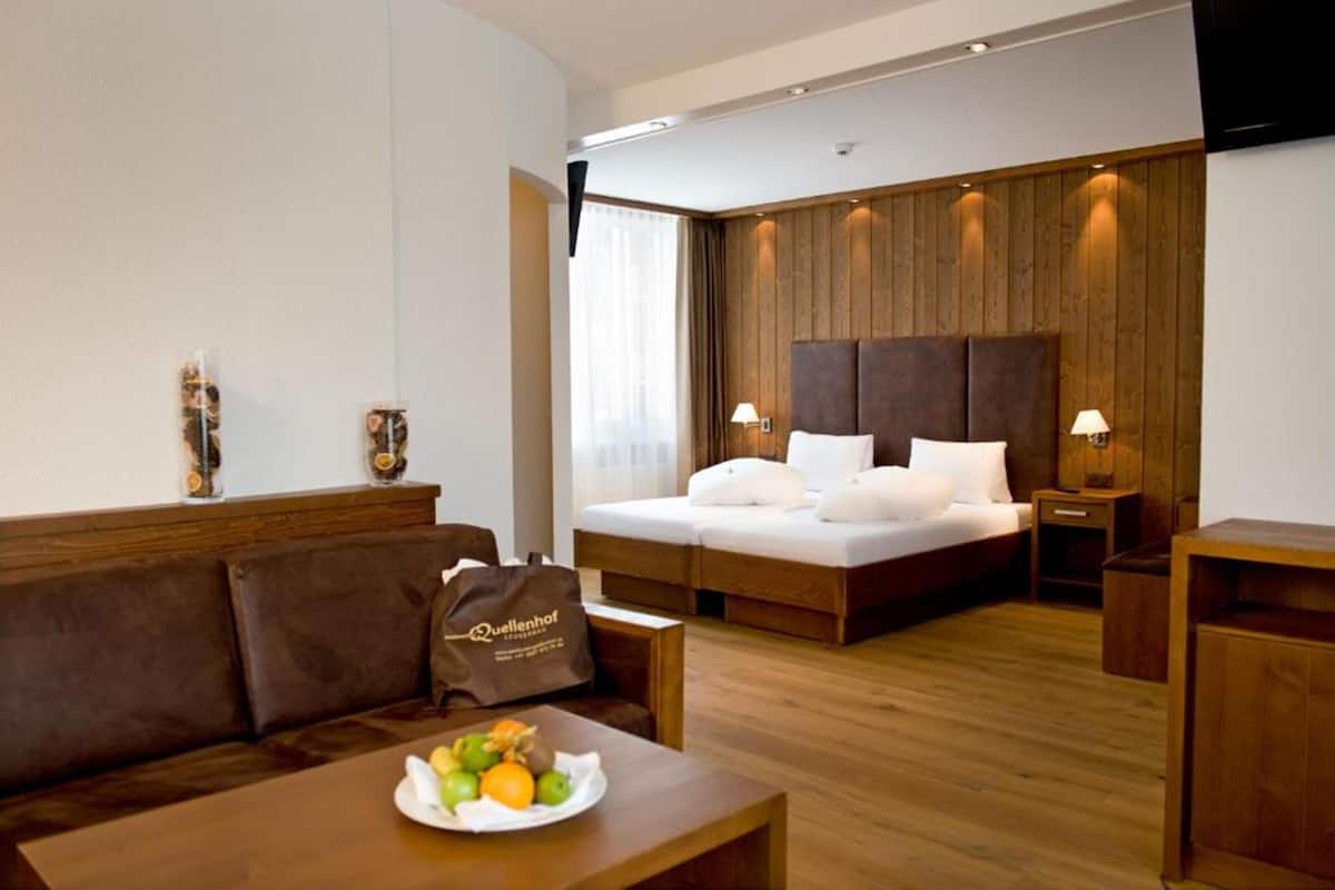 A hotel room with a bed, a wooden headboard, a bedside table with a lamp, a brown leather sofa, and a breakfast tray with fruits.