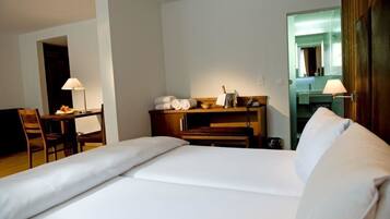 A hotel room with a bed, a desk with a lamp, a chair, and a small table with toiletries.
