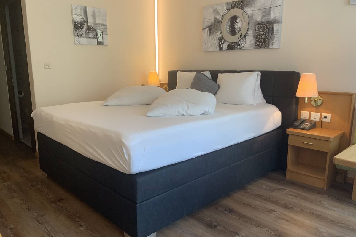 A neatly made bed with white linens and pillows, a bedside table with a lamp, and artwork on the walls.