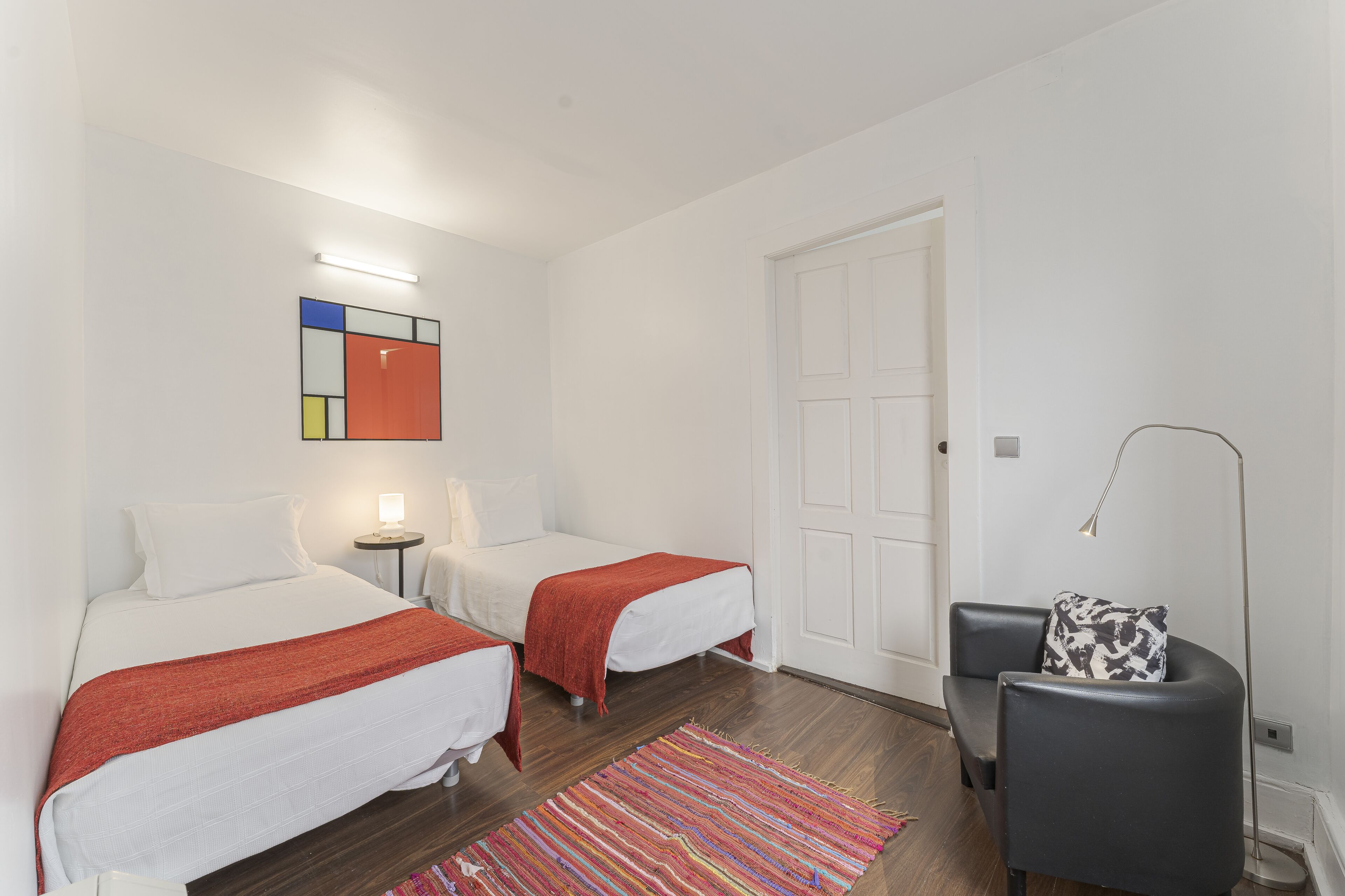 Habitación de hotel con dos camas, una silla, un tapete colorido, una obra de arte en la pared y una puerta.