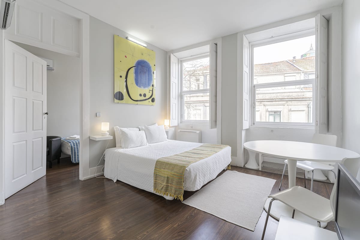 A bedroom with a bed, bedside tables, a desk, and a chair. There is a window with a view of buildings outside.