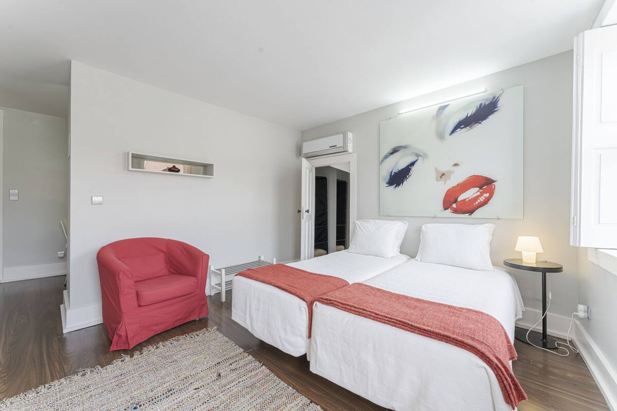 A bedroom with a bed, a red chair, a small table with a lamp, and a painting on the wall.