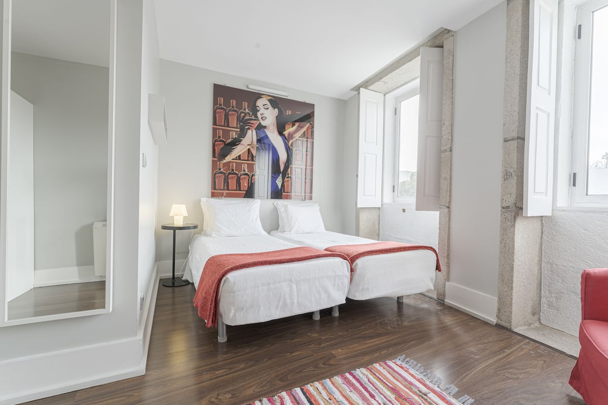 A modern bedroom with two beds, a large window, and a painting on the wall.