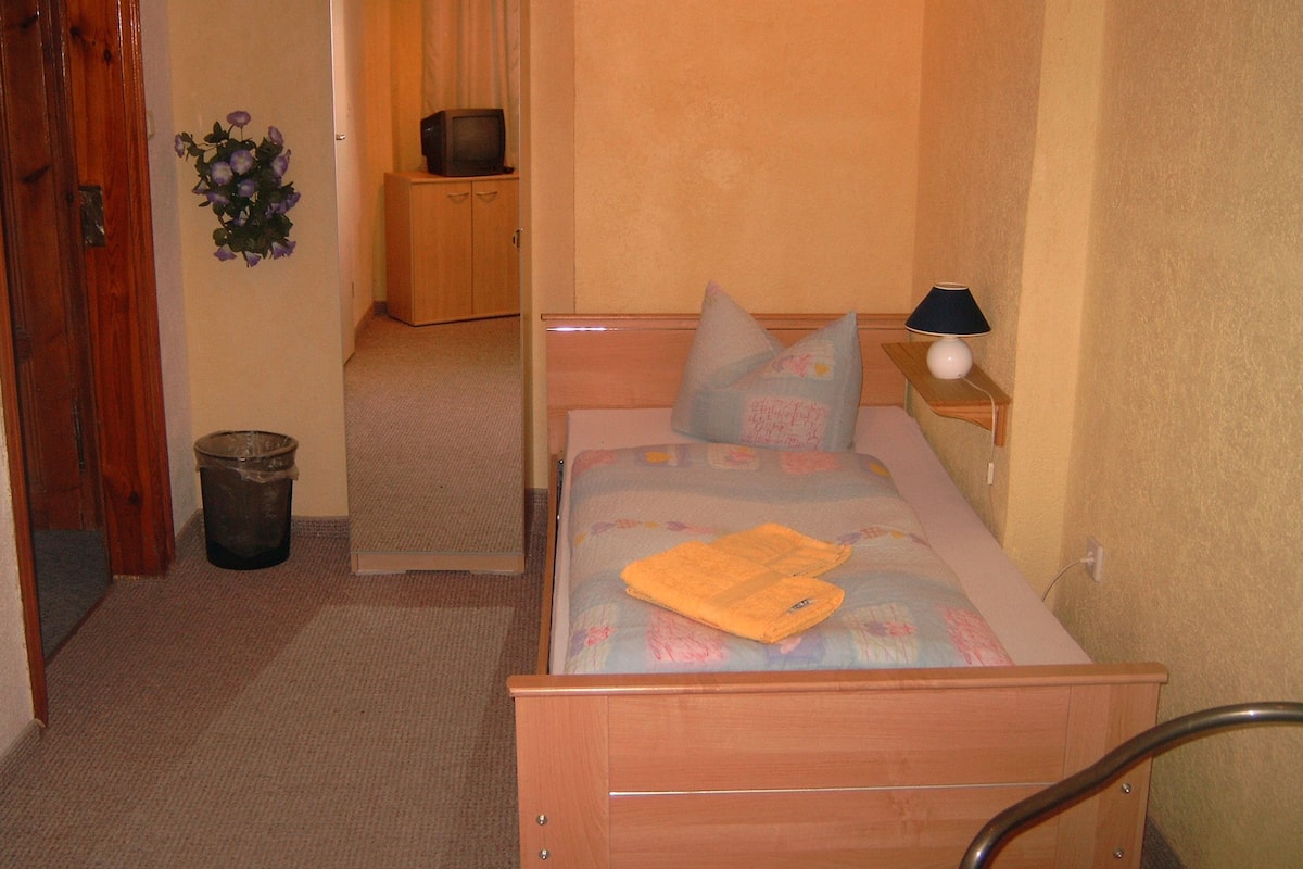 Un dormitorio pequeño y ordenado con cama, almohadas, mesita de noche, lámpara, armario de madera y cubo de basura.