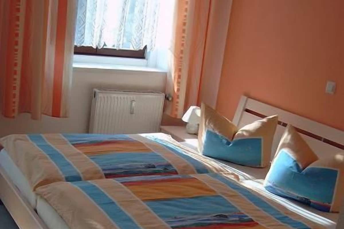 Una cama doble con una colcha a rayas, una mesita de noche con lámpara y una ventana con cortinas.