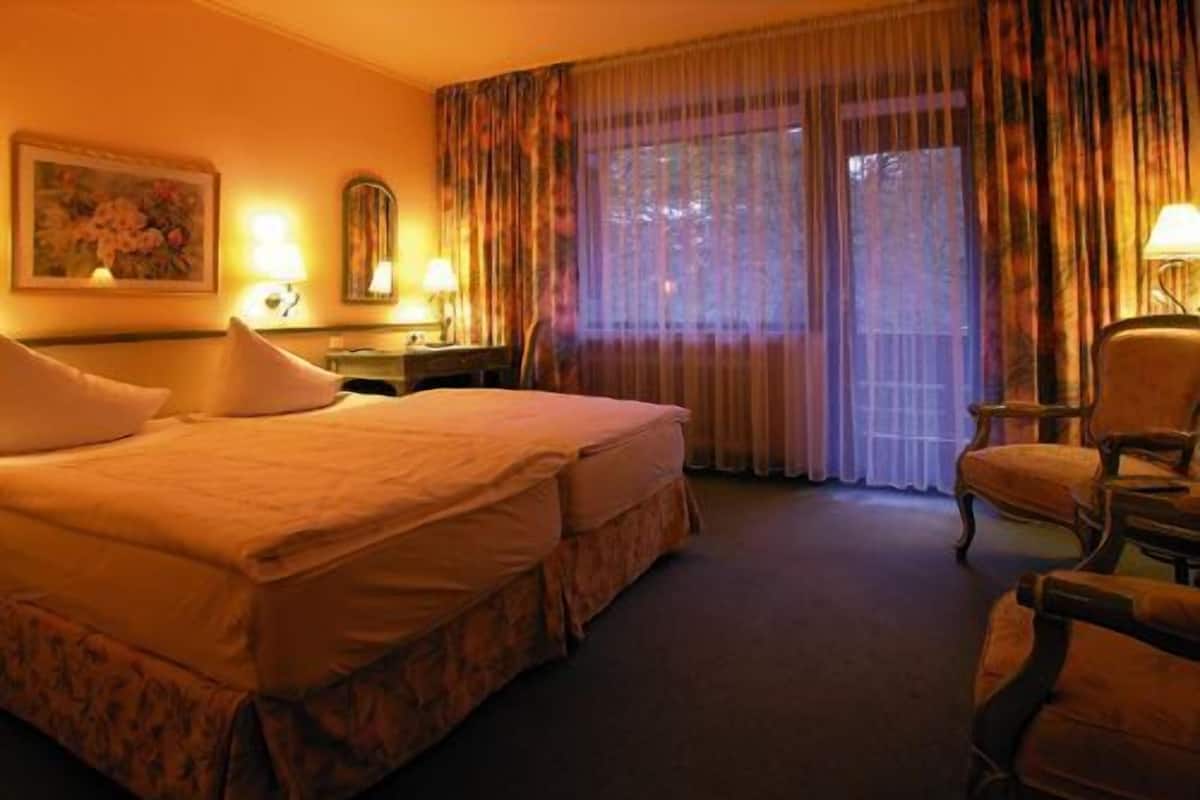 A hotel room with a large bed, two bedside tables with lamps, a chair, and a window with curtains.