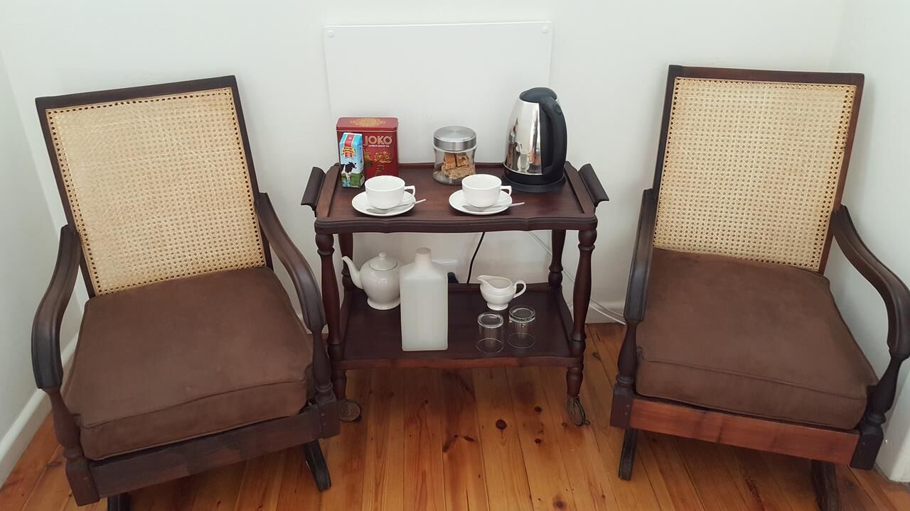 Dos sillones marrones tapizados con respaldos tejidos, una mesa de madera con un juego de té, una caja de té JOKO y una cafetera.