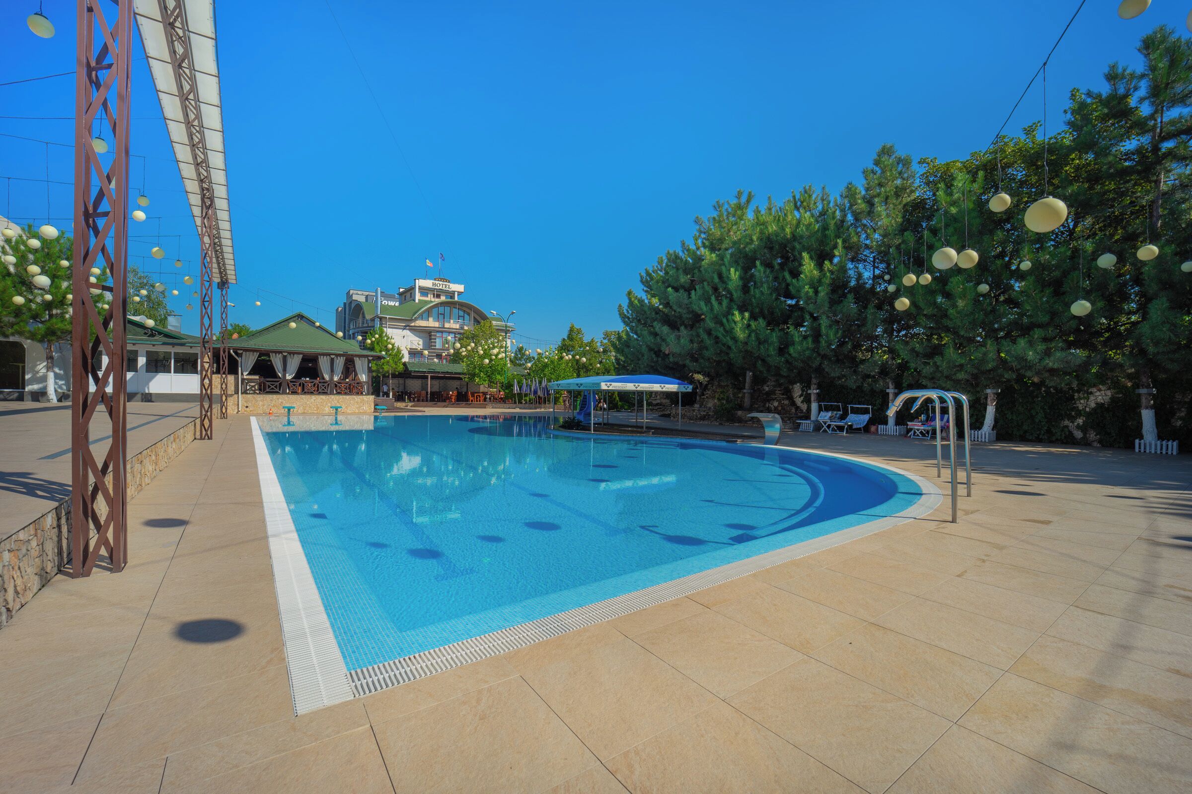A swimming pool with a tiled deck, a covered seating area, and a building with a green roof in the background.