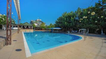 A swimming pool with a tiled deck, a covered seating area, and a building with a green roof in the background.